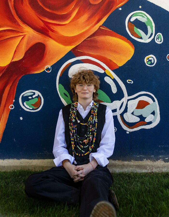 Person sitting on grass wearing colorful bead necklaces in front of a vibrant mural, representing unbelievable luck.