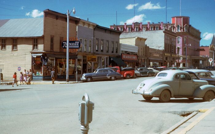 1950s daily life scene with vintage cars and people walking near old storefronts in a small town street.