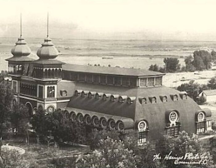 Vintage photo of old architecture featuring detailed windows and unique roof design surrounded by trees and open land.