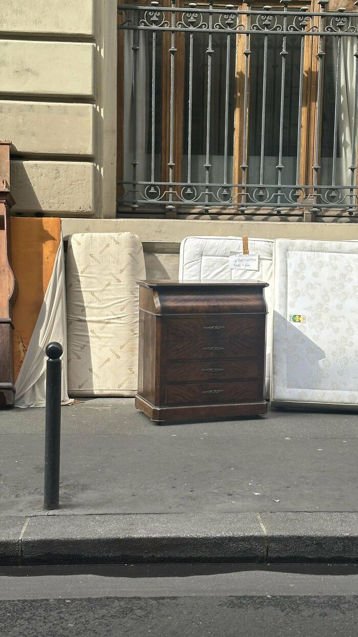 Old wooden chest of drawers and vintage mattresses leaning against a street wall under a decorative metal balcony railing.