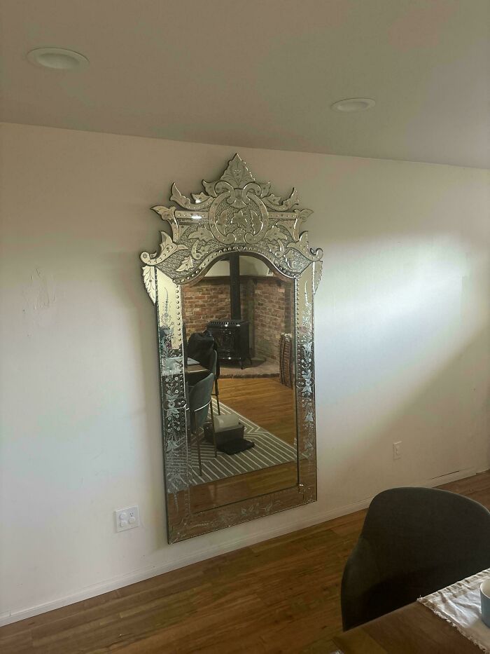 Ornate vintage wall mirror with intricate patterns reflecting a cozy room with wooden floors and a brick fireplace.