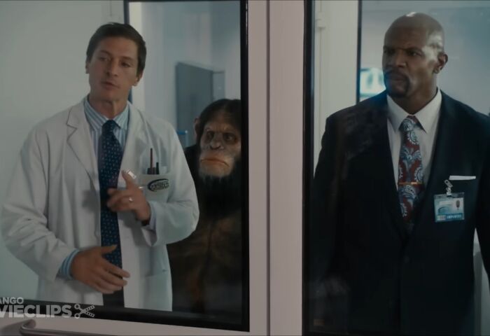 Man in lab coat and man in suit stand by glass with ape character between them in cinematic crimes movie sequel scene.