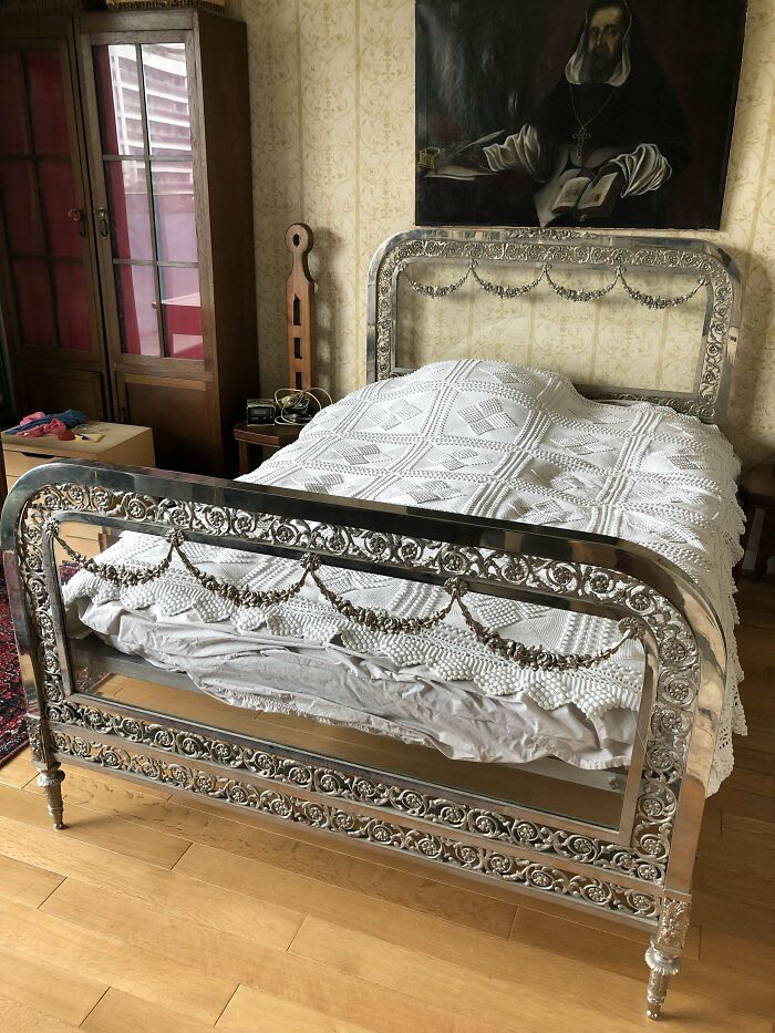 Antique ornate metal bed frame with intricate floral designs and a white crocheted bedspread in a vintage bedroom.