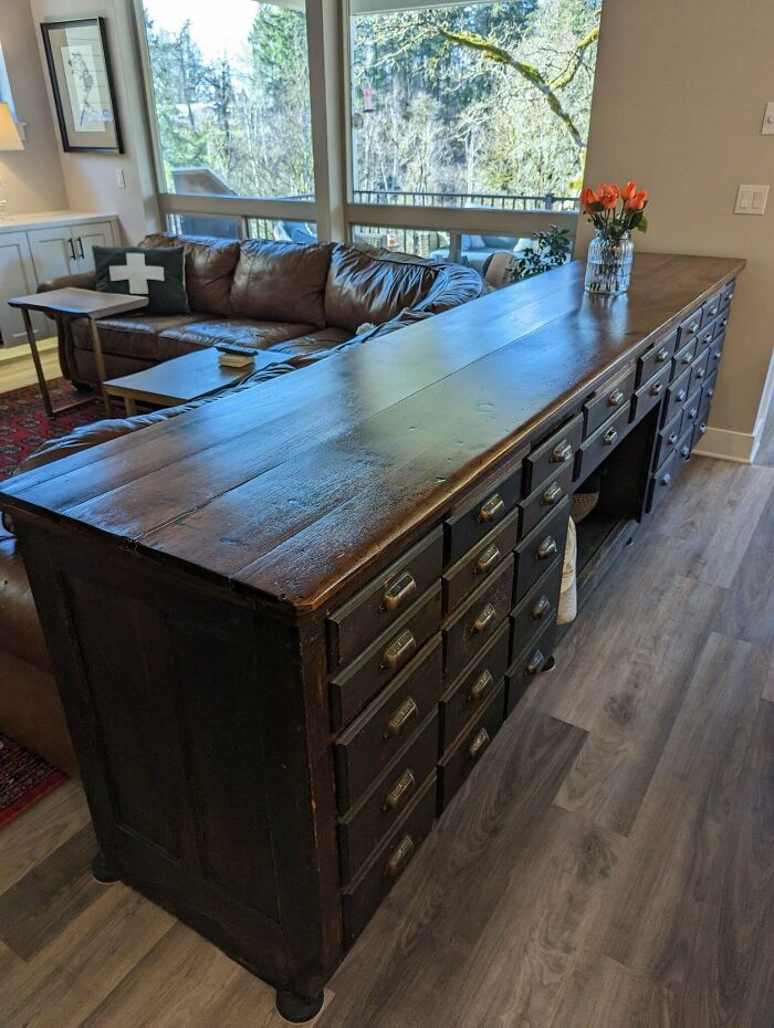 Long wooden antique cabinet with many drawers in a modern living room featuring large windows and leather sofa.