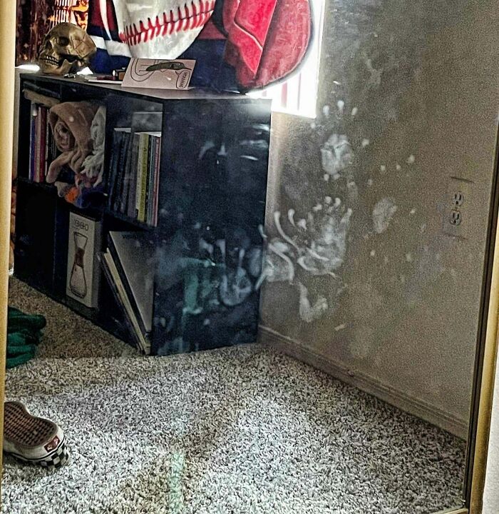 Smudged handprints resembling real-life glitches in the matrix on a mirror beside a shelf with books and decor.