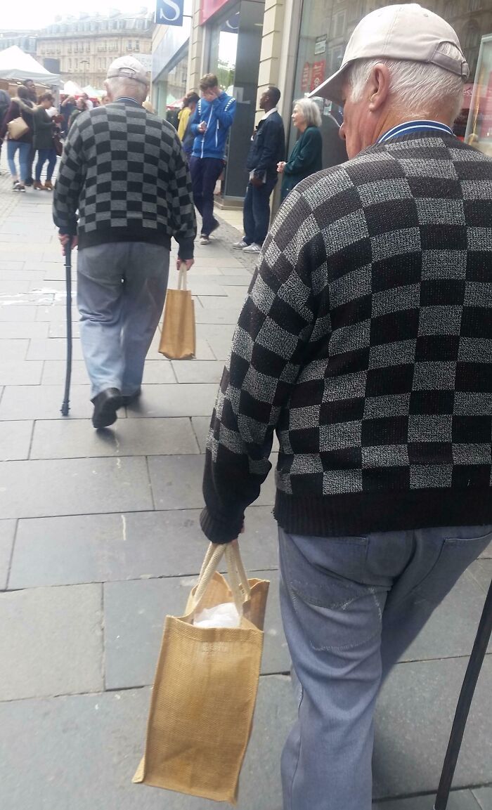 Two elderly men wearing identical checkered sweaters and caps, walking on a busy sidewalk, holding bags and canes, glitch in the matrix.