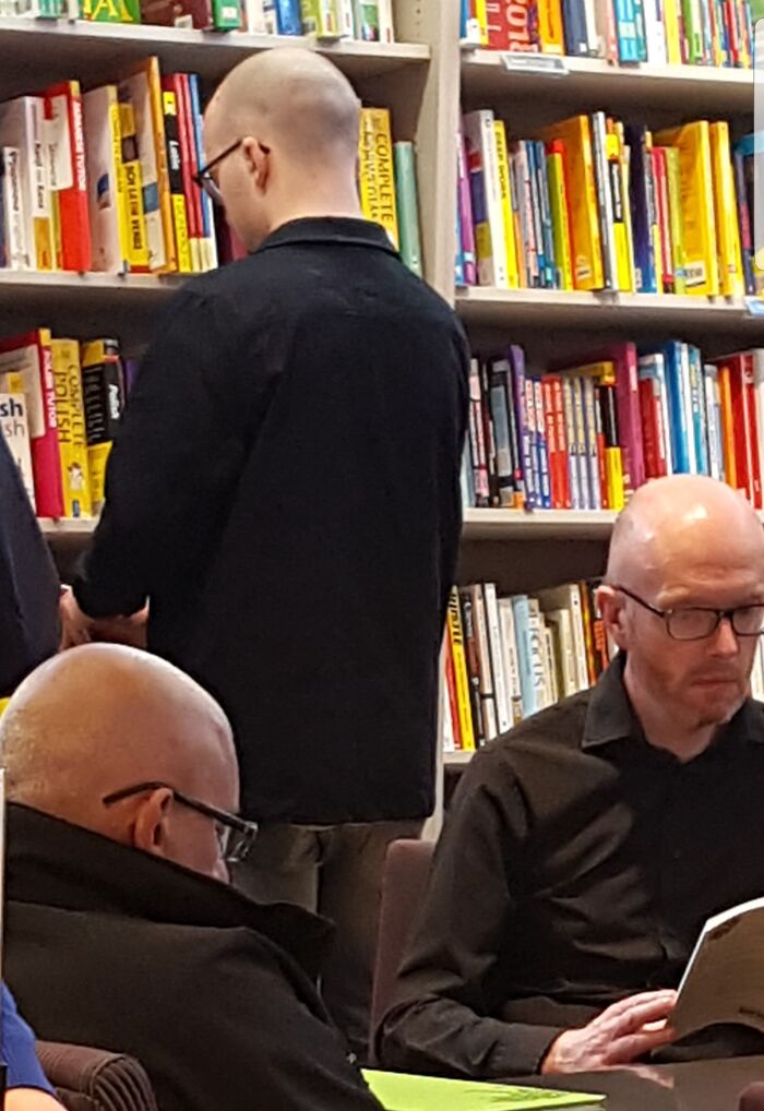 Two identical bald men wearing glasses and black shirts reading books in a library glitch in the matrix moment