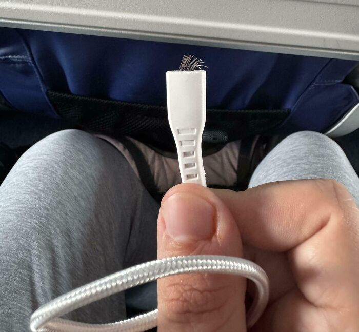 Frayed charging cable held by a person sitting on a plane, highlighting a funny unfortunate situation of forgetting to replace it.