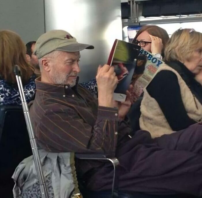 Man sitting unbothered in public, wearing a cap and reading a magazine while others around him are engaged in different activities.