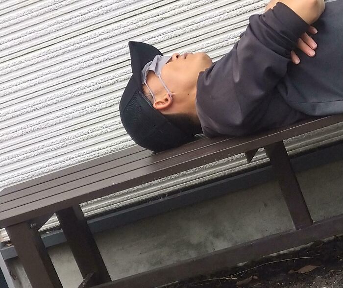 Person lying unbothered on a bench outdoors, wearing a cap and glasses, showing a suspiciously unbothered attitude.