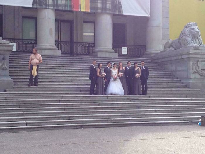 Man shirtless and unbothered standing on steps near formal wedding party posing for photos outside building.