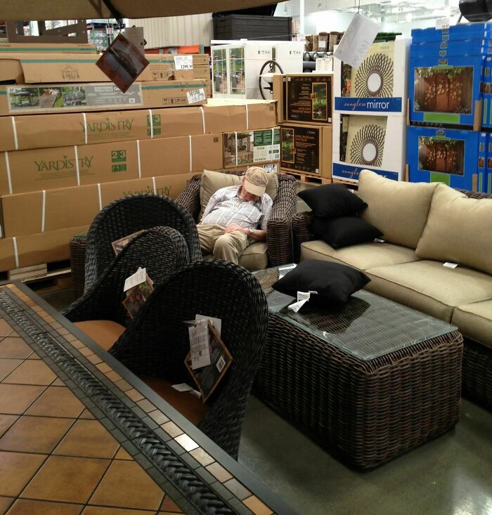 Man sleeping on wicker furniture in a store, suspiciously unbothered amid stacked boxes and displayed items.