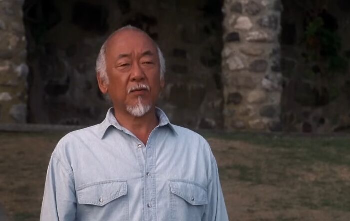 Older man with white hair and beard wearing a light blue shirt standing outdoors near a stone wall in cinematic crimes scene