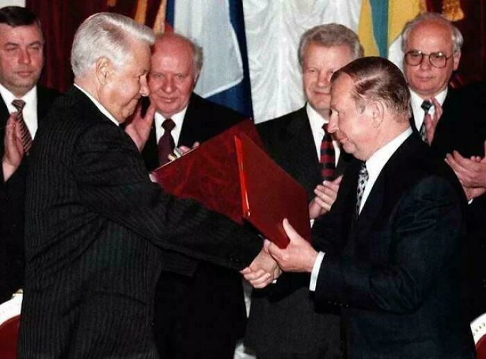 Two men shaking hands and exchanging a red folder in a formal setting with world flags in the background, unusual world facts.