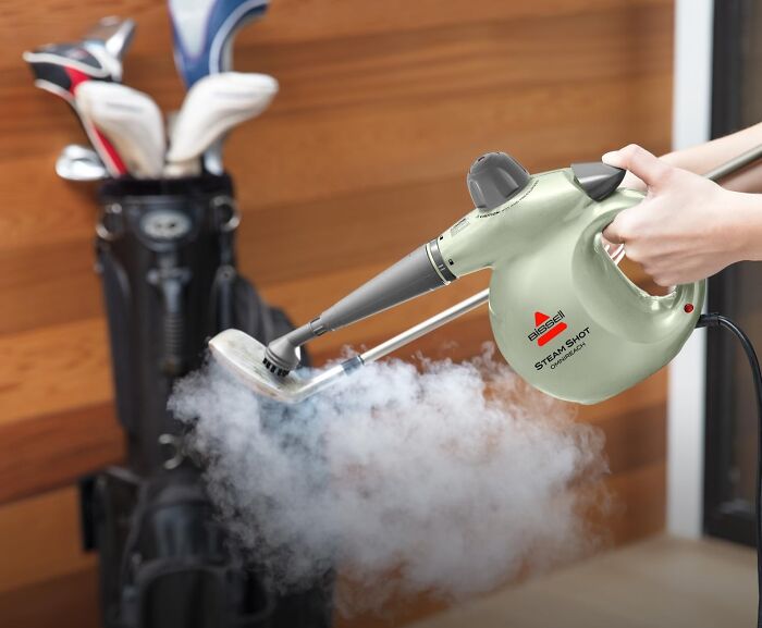 Hand holding a Bissell Steam Shot handheld steamer releasing steam to clean objects in an indoor setting.