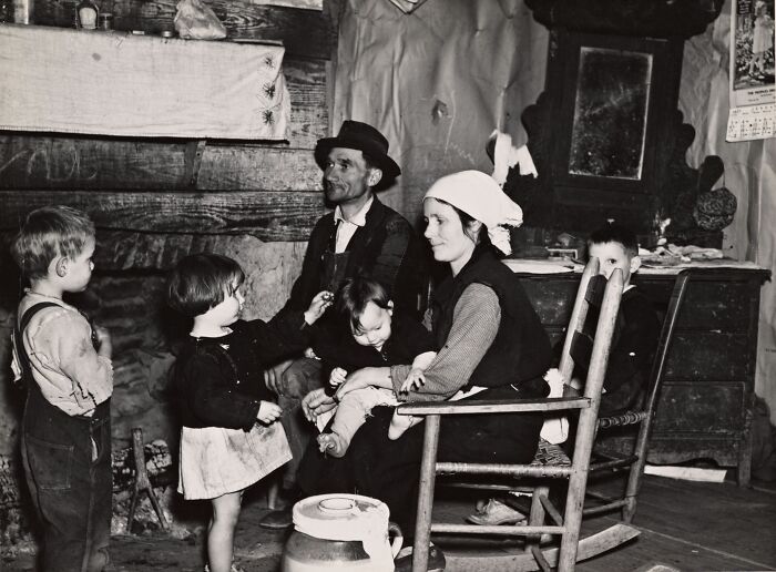 Black and white rare photo of a Great Depression era family gathered inside a rustic home around a fireplace.