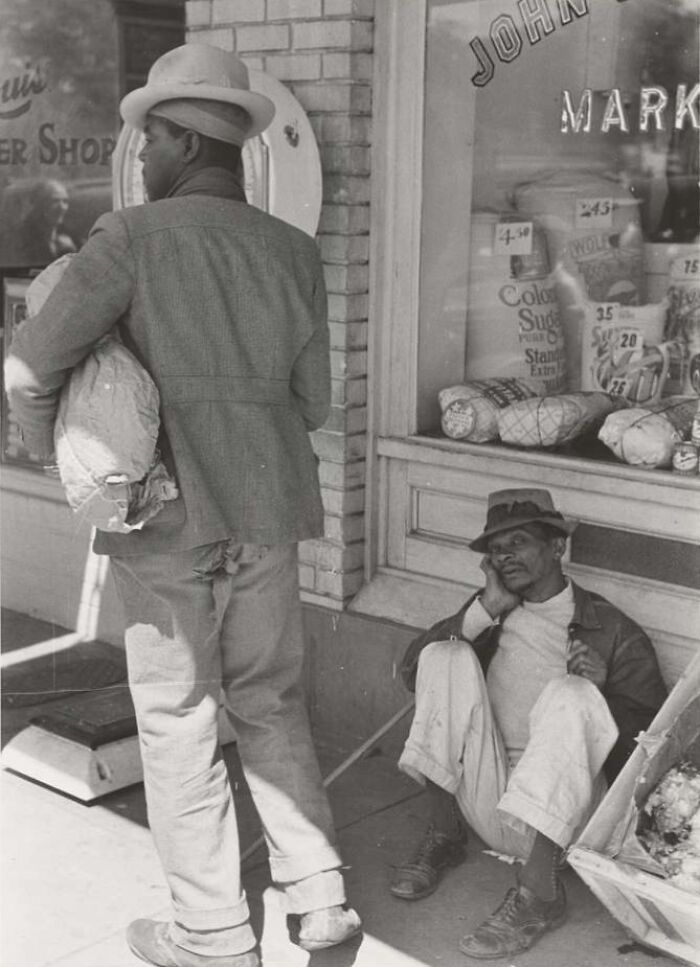 Two men outside a market during the Great Depression era, one sitting on the ground and the other standing holding a sack.