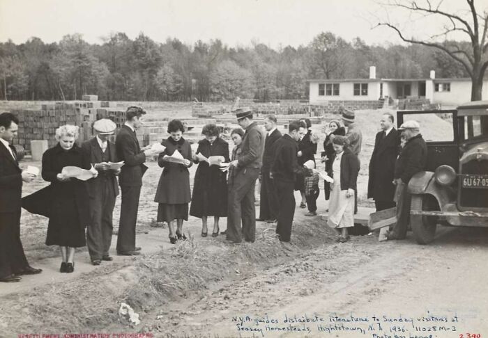 Group of people at an outdoor event during the Great Depression era, receiving literature on a dirt road near a vintage car.