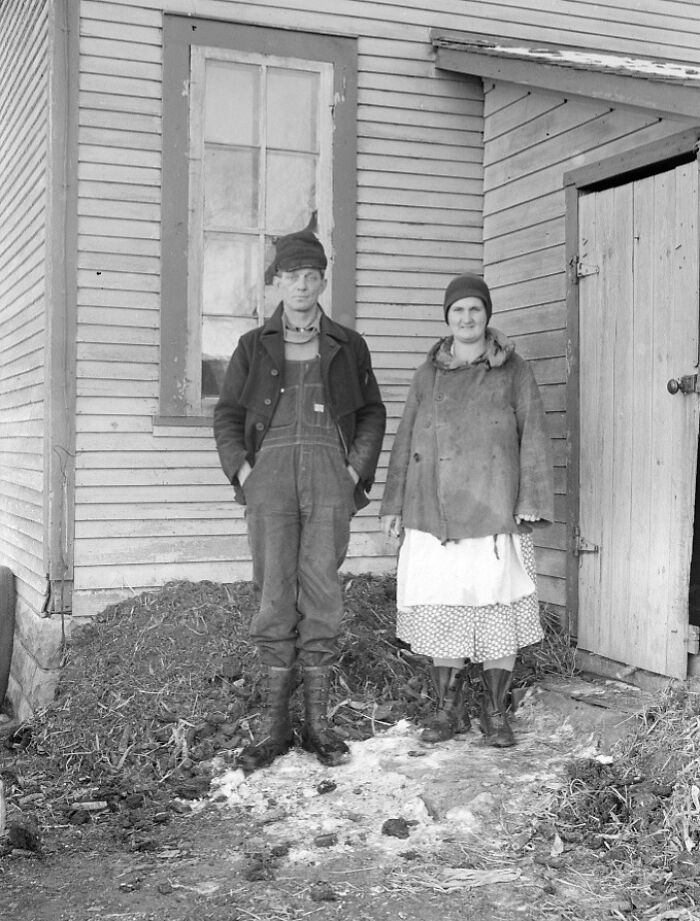 Black and white photo of a Great Depression era couple standing outside a wooden house, dressed in worn clothes and boots.