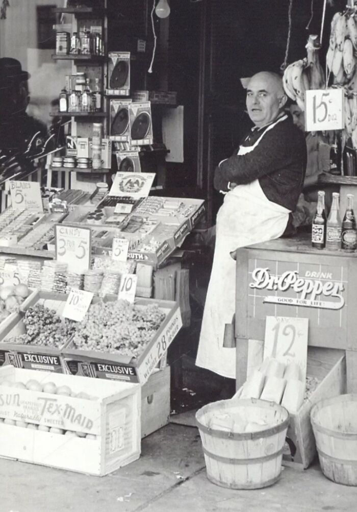 Man in apron standing in a grocery store surrounded by fruits, candies, and vintage Depression era prices and products.