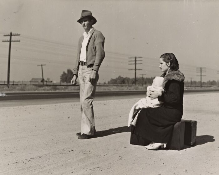 Man standing and woman sitting with baby on suitcase by roadside during Great Depression era in rare photo.