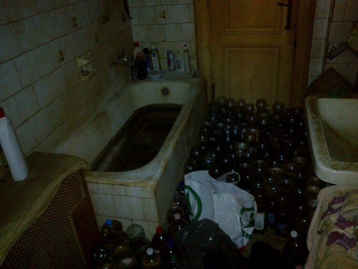 Dimly lit, cluttered bathroom with numerous bottles and jars scattered around a dirty bathtub and sink.