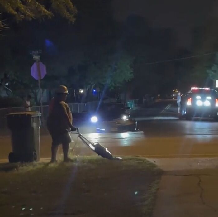 Person mowing lawn at night with headlights and police car lights in the background showing suspiciously unbothered attitude.