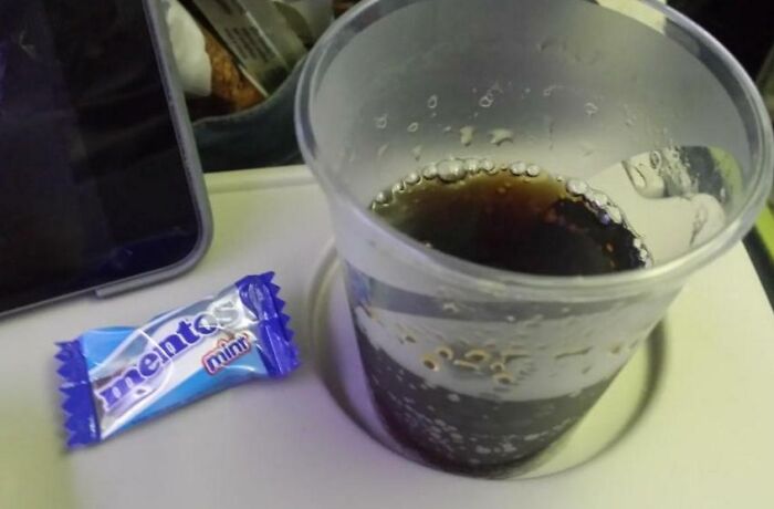 Clear plastic cup with soda on airplane tray table next to Mentos mint candy, showing suspiciously unbothered details.