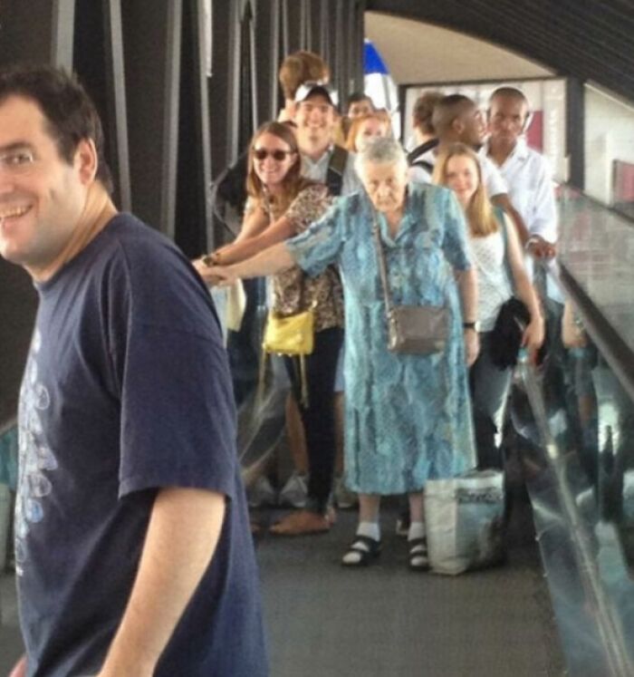 A smiling man blocks a group of people on a moving walkway, showing suspiciously unbothered attitudes in public.