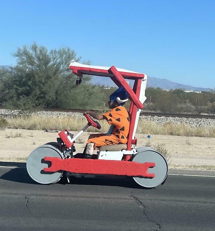 Person dressed in a Flintstones costume driving a homemade car, suspiciously unbothered while on the road.