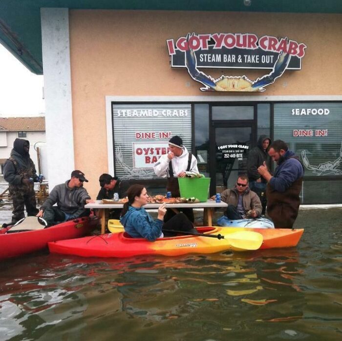 People suspiciously unbothered eating seafood at a flooded restaurant while sitting in kayaks and chairs in rising water.
