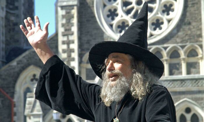 Man dressed as a wizard with a large black hat gestures outdoors near a medieval stone building with circular window, unusual world facts.