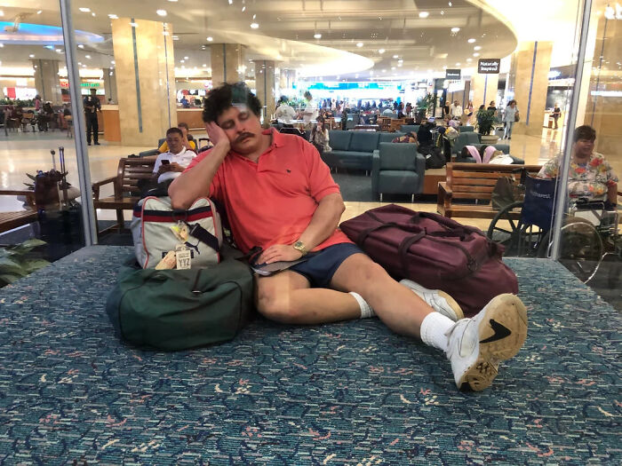 Man in a coral shirt resting on carpeted floor surrounded by bags at a busy airport lounge, unusual world facts setting.