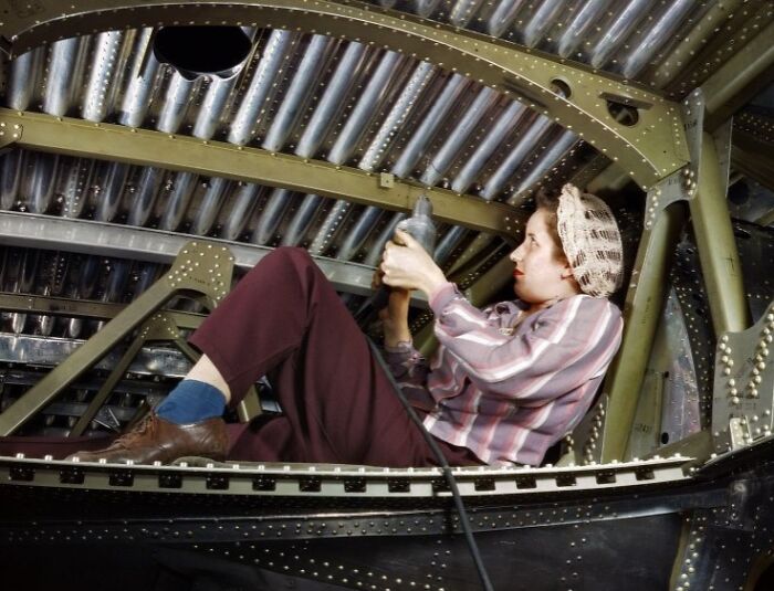Woman at work during WWII wearing a headscarf, operating machinery inside an aircraft undercarriage structure.
