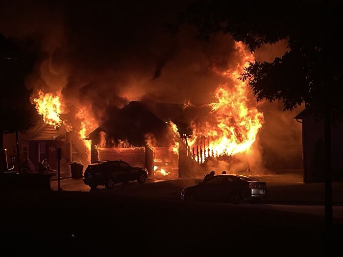 House engulfed in flames at night with cars nearby, illustrating dramatic neighbor tales in a residential wooded area.