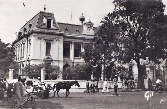 Black and white photo of daily life across the globe in the 50s showing people, horse cart, and cyclists near a historic building.