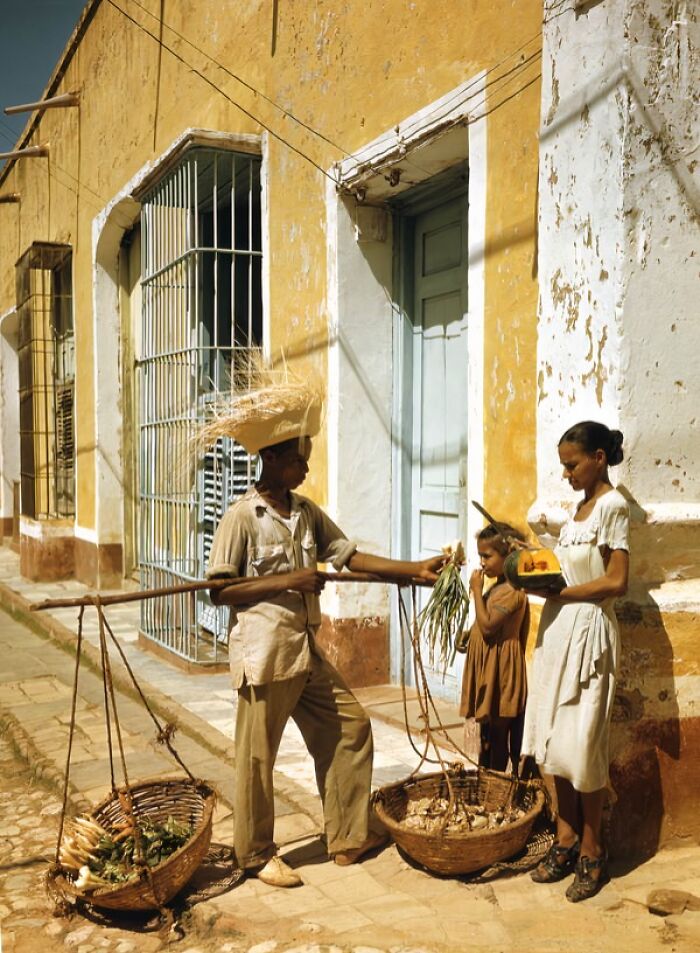 Man selling vegetables to woman and child on a street, depicting daily life across the globe in the 50s.