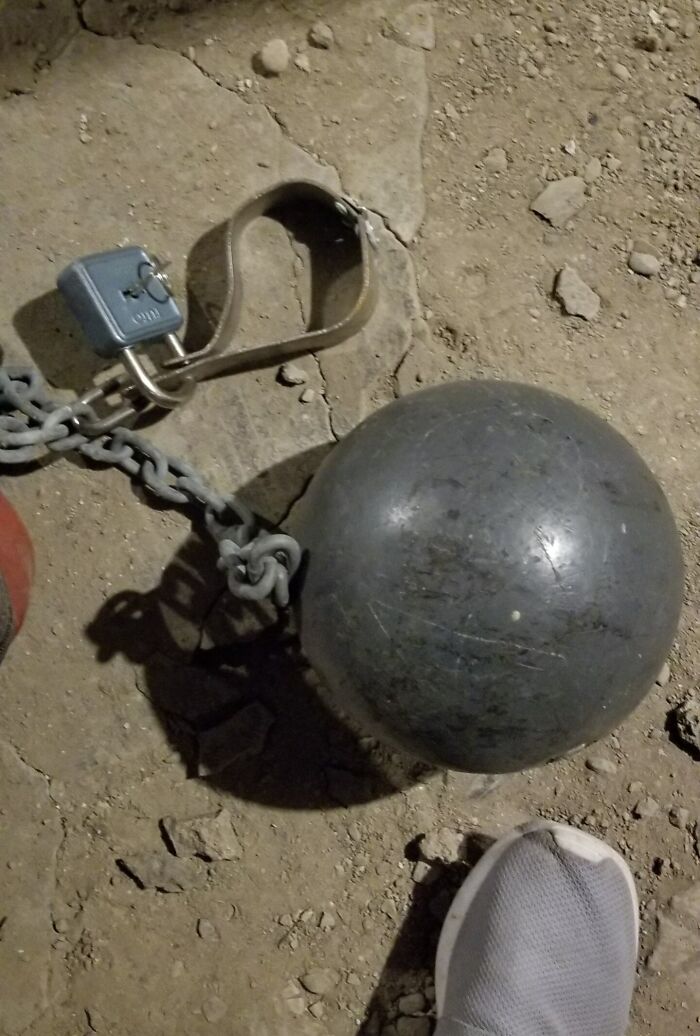 Heavy iron ball with chain and lock on dusty floor next to a sneaker, an unusual find in an old home.