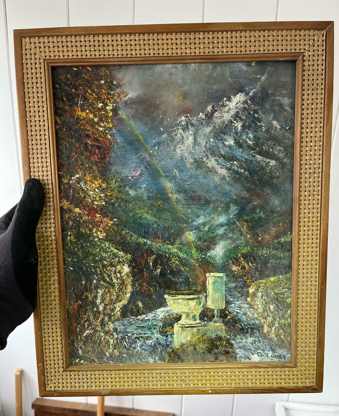 Framed painting of a mountain landscape with a rainbow and river, held by a gloved hand inside an old home.