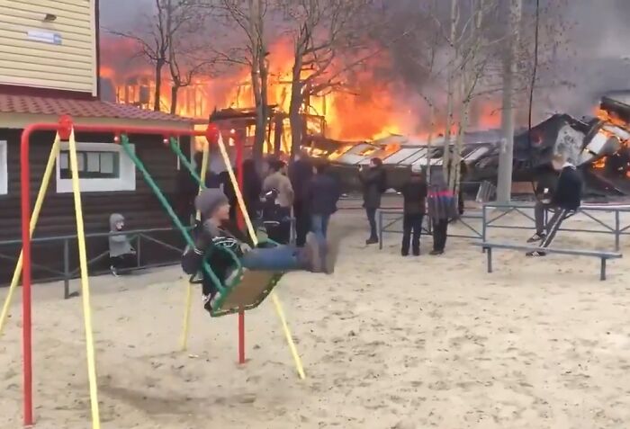People on swings and nearby appear suspiciously unbothered as a large fire engulfs a building in the background.