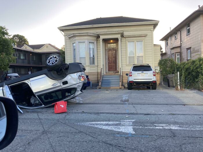 Overturned delivery car on residential street with a red package on the ground showing unfortunate delivery mishap.