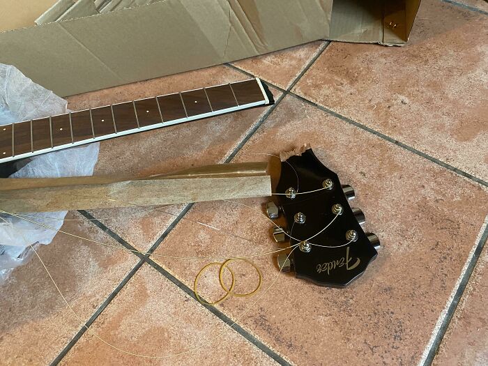 Broken Fender guitar with detached neck lying on tiled floor next to a cardboard package, showing delivery mishap.