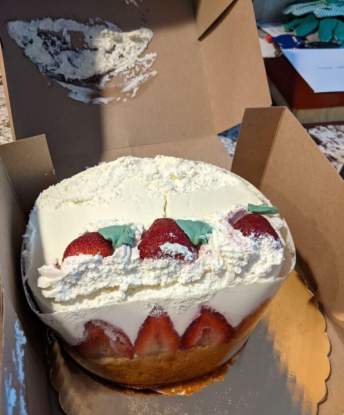 Partially damaged strawberry cake with whipped cream inside a cardboard delivery box, showing a delivery mishap.