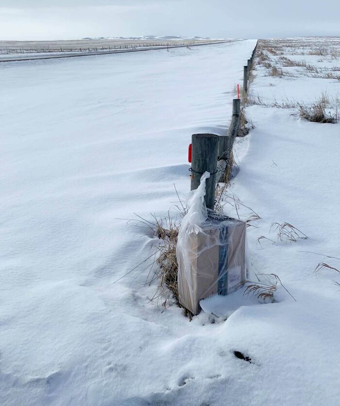 Package delivery wrapped in plastic and partially buried in snow along a rural fence in a remote snowy area.