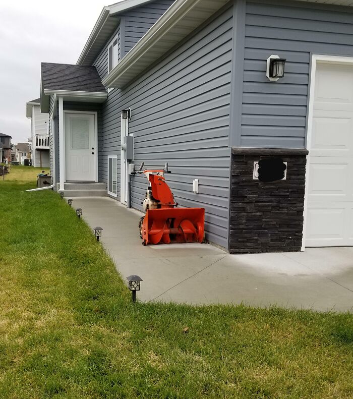 Side view of a house with a snowblower parked on the walkway near the door and green grass on the side.