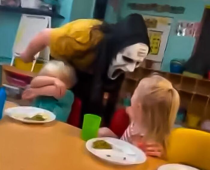 Person wearing scary mask holding child near surprised kid at table, illustrating consequences of stupidity and lost jobs.