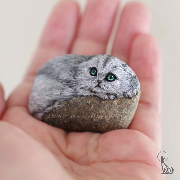 Small stone painted by Japanese artist into an adorable animal with detailed fur and bright green eyes held in a hand.