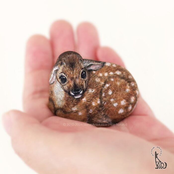 A small hand holding a stone painted by a Japanese artist to look like an adorable sleeping fawn.