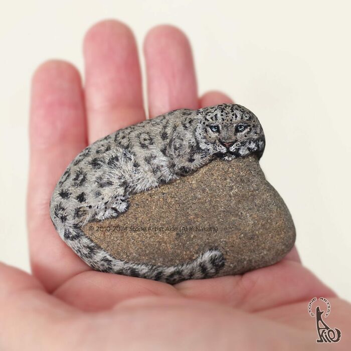 Hand holding a stone painted by a Japanese artist to resemble an adorable resting snow leopard animal.