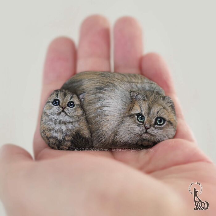 Hand holding stones painted by Japanese artist to resemble adorable animals with detailed fur and expressive eyes.